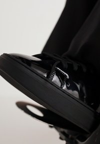 Μαύρο παπούτσι σνίκερ από γυαλιστερό δέρμα με λείο υφή, στρογγυλεμένη μύτη, μαύρους κορδόνες και παχύ μαύρο ελαστικό πάτο. Η κοντινή λήψη τονίζει τη λάμψη.