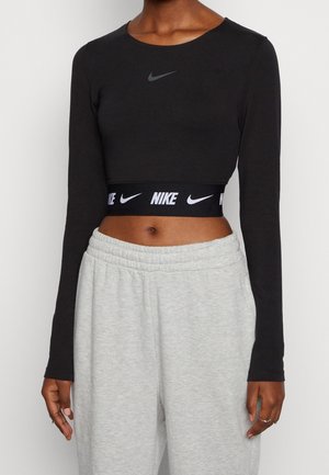 Femme portant un crop top noir à manches longues Nike avec un logo gris sur la ceinture et un pantalon de survêtement gris clair, debout sur un fond blanc.