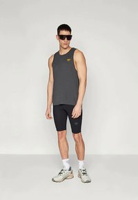 Siva bezrukavna majica s žutim logotipom, uparena s crnim uskim kratkim hlačama i svijetlosivim sportskim cipelama. Glatka tkanina, ležeran dizajn.