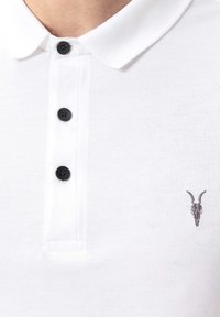 Polo bianca con bottoni neri e un piccolo teschio di animale scuro ricamato con corna curve sul petto.