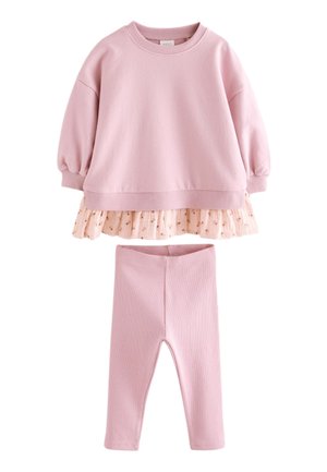 Småbarnspige med skulderlangt mørkt hår iført en pink oversize sweater, blomsteret nederdel, pink leggings og cremefarvede højtop sneakers.