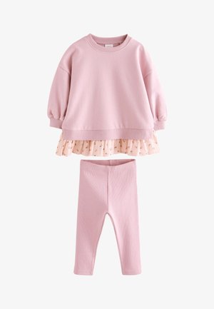 Dziewczynka z ciemnymi, sięgającymi do ramion włosami, ubrana w różowy, oversize'owy sweter, kwiecistą spódnicę, różowe legginsy i kremowe sneakersy za kostkę.