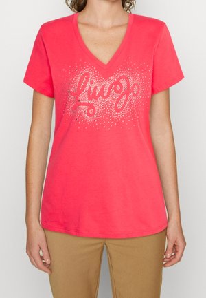 Kvinde iført en knaldpink V-hals T-shirt med sølvprikket tekstdesign og beige bukser, stående mod en ensfarvet baggrund.