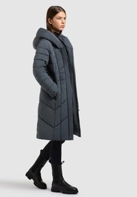 Odebeljen sivi plašč z visokim ovratnikom in šivi v obliki chevrona; nošen preko črne majice, kombiniran s črnimi usnjenimi škornji do sredine meč.