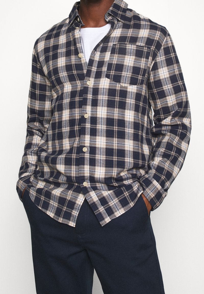 Koszula w niebiesko-beżową kratkę z długimi rękawami, z przodu zapinana na guziki, z kieszenią na piersi. Nosi się ją na białej koszulce, zestawioną z ciemnoniebieskimi spodniami.