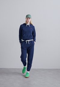 Navy blue athletic hoodie with white drawstrings and pocket, paired with matching joggers. Model wears green sneakers and a green cap.