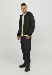 Czarna kurtka zapinana na zamek, wykonana z gładkiego materiału, zestawiona z luźnymi spodniami cargo z bocznymi kieszeniami. Czarne buty dopełniają outfit.