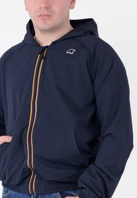 Giacca a vento blu navy con cappuccio in materiale leggero, dotata di dettagli colorati sulle cerniere e doppie tasche anteriori, con polsini e orlo elastici.
