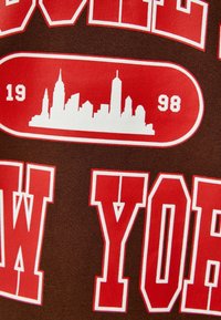 Brunt tyg med vita och röda grafik. Innehåller "NEW YORK" i fet stil och en stadssilhuett inom en rundad rektangel.