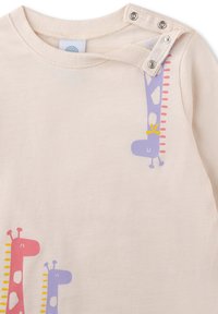 Helle beige Baumwollbluse mit Giraffenmotiven in Pink und Lila, ausgestattet mit Druckknöpfen an der Schulter für ein einfaches An- und Ausziehen.