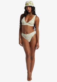 Frau in grün-weißem gemustertem Bikini und passendem Bucket Hat, steht mit entspannter Haltung vor einem weißen Hintergrund.