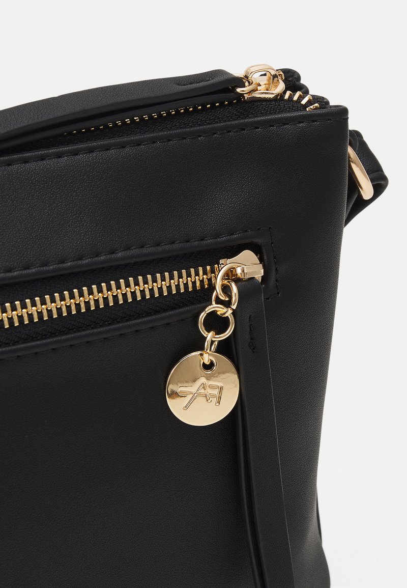Bolso de cuero sintético negro con cremallera y herrajes dorados, con un charm decorativo dorado adjunto al tirador de la cremallera.
