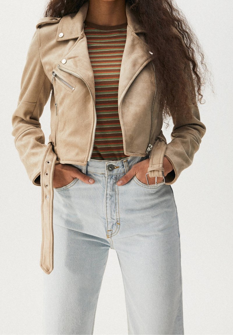 Femme portant une veste biker courte en daim beige avec poches zippées et ceinture, un top rayé marron et vert, et un jean taille haute bleu clair.