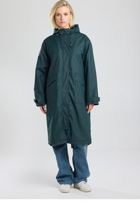 Longue veste imperméable vert foncé avec capuche, fermeture à pression, deux poches frontales et coupe ample, portée sur un jean à jambes larges et des baskets.