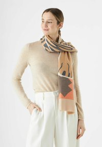 Foulard beige et marron à motifs avec des designs abstraits, fabriqué en matière douce, drapé autour du cou, associé à un haut clair et un pantalon blanc.