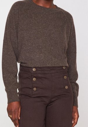 Personne portant un pull en maille marron tacheté et un pantalon taille haute marron foncé avec deux rangées de boutons en laiton à l’avant.