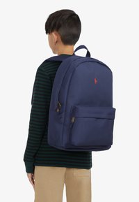 Tmavě modrý batoh z odolné tkaniny, který má přední kapsu a výrazné logo. Design zahrnuje polstrované nastavitelná ramenní popruhy a strukturovaný tvar.