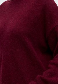Gros plan d'une personne portant un pull doux et duveteux rouge foncé à manches longues sur un fond uni.