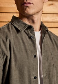 Man wearing an olive green corduroy button-up shirt over a white t-shirt, shown from the neck to mid-chest against a wooden background.