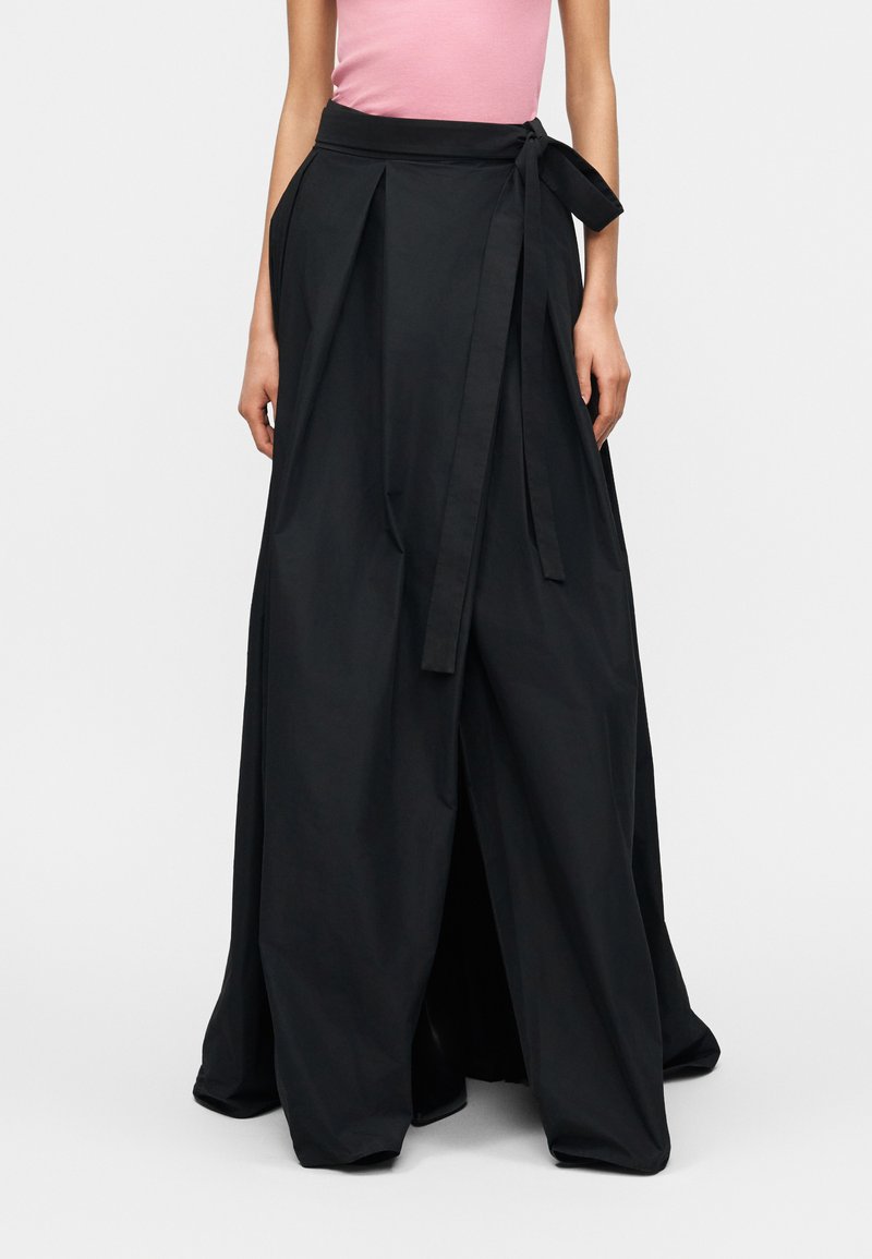 Γυναίκα που φοράει μακριά, φαρδιά ασύμμετρη μαύρη φούστα με ζώνη και ροζ αμάνικο τοπ μέσα στη μέση.