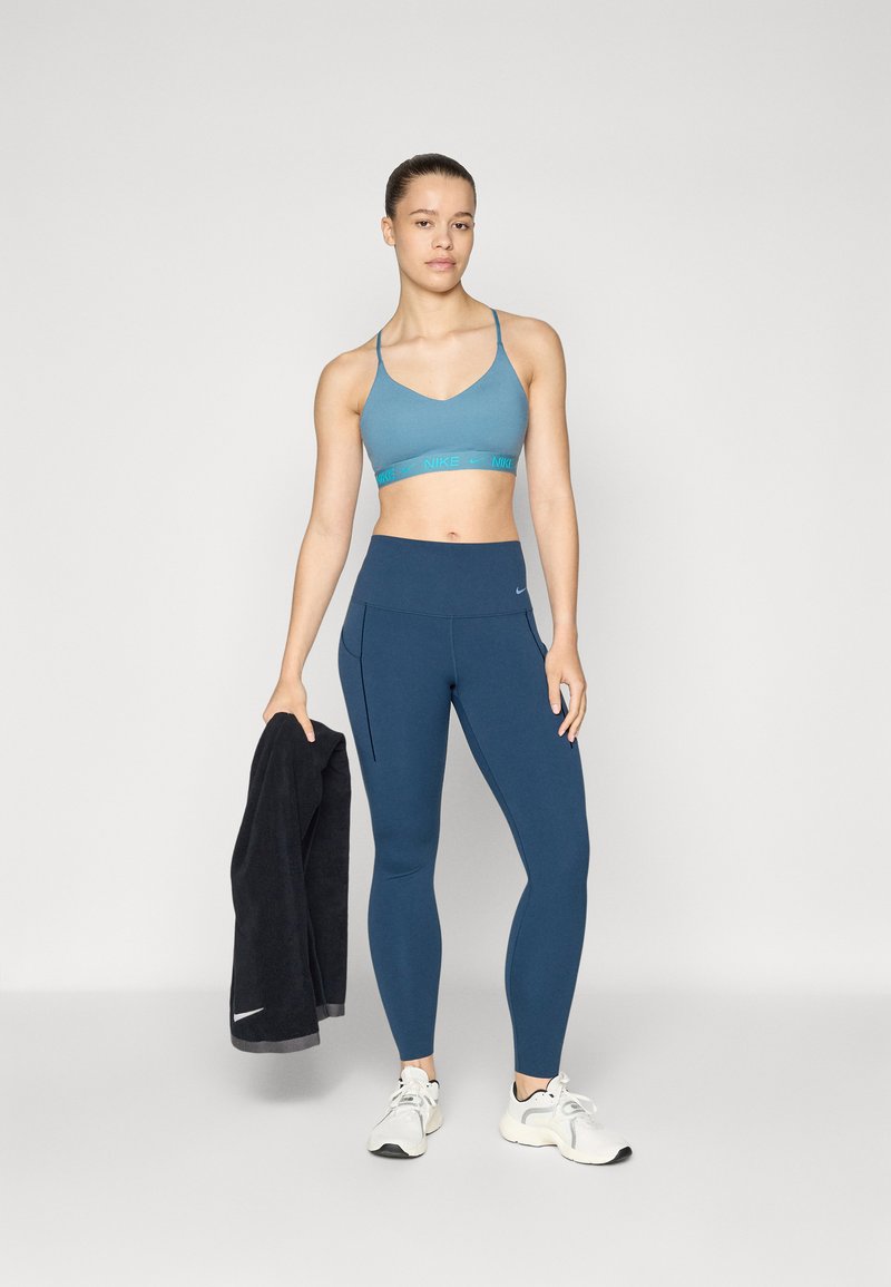 Sujetador deportivo azul con detalle de logo, leggings azul de cintura alta y zapatillas grises. Toalla oscura colgada del brazo, textura de tela suave.