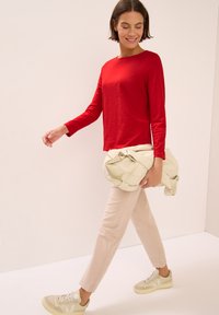 Camicia rossa a maniche lunghe, pantaloni beige e borsa crema. Il modello indossa scarpe da ginnastica chiare. Design semplice, texture liscia e vestibilità casual.