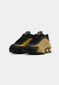 Μαύρα και χρυσά Nike Shox παπούτσια με μαύρες κορδόνες, διατρητές λεπτομέρειες και χαρακτηριστικούς στυλοβάτες στην φτέρνα.