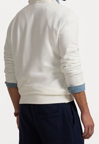 Man som bär en vit långärmad tröja med blå uppvikt skjortärmar och marinblå byxor, står med ryggen vänd mot kameran.