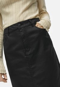 Jupe en denim noir à la texture lisse, dotée de poches avant, d'une fermeture à boutons, et d'une silhouette taille haute. Le pull présente un motif tricoté.