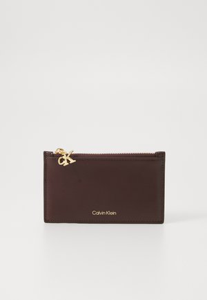 Brown leather card holder with a gold zipper pull featuring "CK" logo, and embossed "Calvin Klein" text. Smooth texture, minimal design.