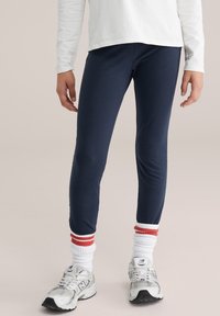 Sötétkék leggings testhezálló fazonban, puha anyagból, fehér zoknival, amelyen piros és tengerészkék csíkok vannak, valamint szürke sportcipővel párosítva.