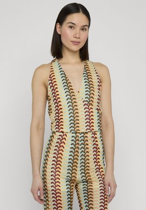 Femme aux cheveux raides et foncés portant une combinaison colorée sans manches à motifs avec un décolleté en V profond sur un fond uni clair.