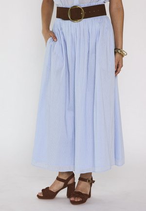 Femme portant une jupe plissée bleu clair avec une large ceinture marron et des sandales marron à talons assorties, mains reposant sur les côtés.