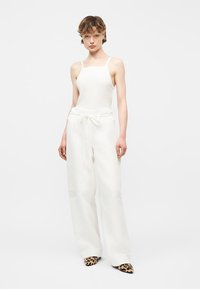 Personne portant un haut blanc sans manches et un pantalon blanc à jambes larges avec cordon de serrage, associé à des chaussures pointues à imprimé léopard, debout devant un fond blanc.