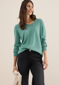 Ljus teal, långärmad tröja med V-ringning och ribbade ärmslut, kombinerad med svarta byxor. Personen håller i en beige, texturerad föremål.