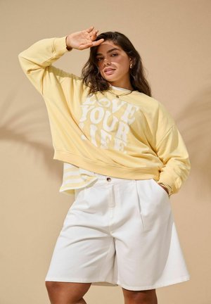 Kobieta o długich włosach, ubrana w żółtą bluzę z napisem „LOVE YOUR LIFE” oraz białe szorty, pozująca z ręką przy czole na beżowym tle.