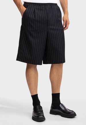 Mand iført sorte knælange shorts med nålestriber, sorte sokker og sorte lædersko uden snørebånd foran en ensfarvet baggrund.