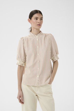 Nő krémszínű blúzban, amelyen függőleges piros csíkok vannak, púposított rövid ujjakkal és bézs nadrágban, egy kézzel a zsebében áll.