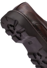 Zapato de cuero marrón texturizado con una suela de goma negra robusta que presenta un patrón de agarre y un logotipo en relieve en el lado.