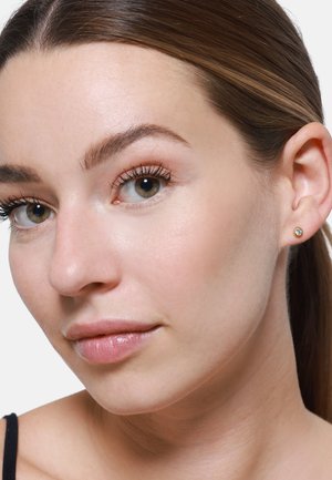 Close-up van een gezicht van een persoon, met een soepele huid, lichte make-up en kleine gouden studs met heldere kristallen. Het haar is naar achteren gepind.