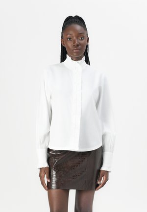 Camisa blanca de botones con cuello de volante y mangas largas, combinada con una falda ajustada de piel de cocodrilo marrón, que muestra textura y contraste.