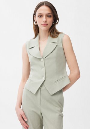 Junge Frau mit hellbraunen Haaren, die eine ärmellose, hellgrüne, maßgeschneiderte Weste und dazu passende hoch taillierte Hosen trägt, vor weißem Hintergrund stehend.