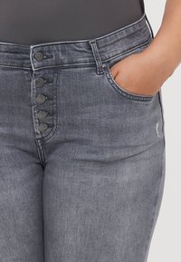 Dark grey jeans in denim fabric featuring five buttons at the waist, side pockets, and minimal distressing on the right leg.