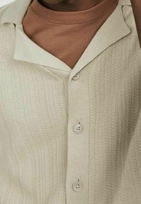 Beige stickad kavaj med en texturerad yta, spetskrage och tre knappar. Buren över en brun polotröja.