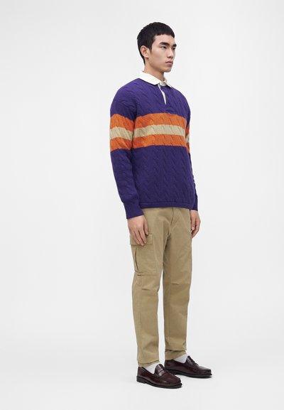 Homme debout portant un pull en maille torsadée violet avec des rayures beige et orange, un pantalon cargo beige, des chaussettes blanches et des mocassins marron foncé.