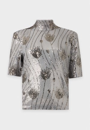 Une femme porte un haut gris à sequins, une jupe midi beige avec un ourlet en dentelle, des bottes marron et un sac à main bordeaux, sur un fond uni.