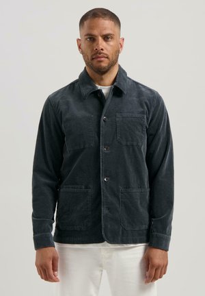 Man in een donkergrijze corduroy button-up jas met drie voorzakken over een wit shirt en witte broek, staande tegen een effen achtergrond.
