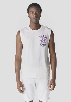 Joven con camiseta sin mangas blanca con texto "Osaka Beach" y pantalones cortos blancos rasgados, posando contra un fondo claro y liso.