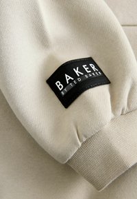 Tessuto beige chiaro con una superficie testurizzata, caratterizzato da un'etichetta intrecciata nera che riporta la scritta "BAKER by TED BAKER" in lettere bianche sulla manica.