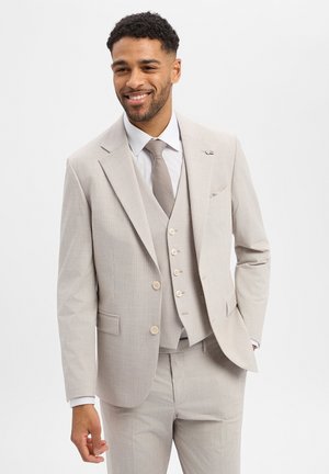 Mann lächelnd, trägt einen hellbeigen Dreiteiler mit weißem Hemd und gemustertem beigen Krawatte, steht vor einem schlichten weißen Hintergrund.